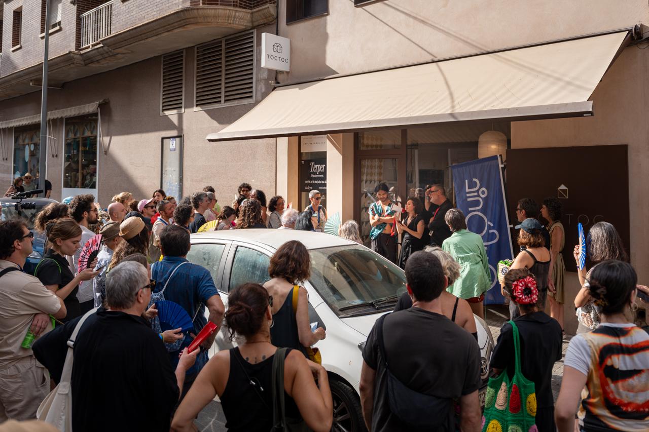 A crowd gathered on the street outside TOCTOC in Amposta, fanning themselves, the Displacement poster visible in the doorway