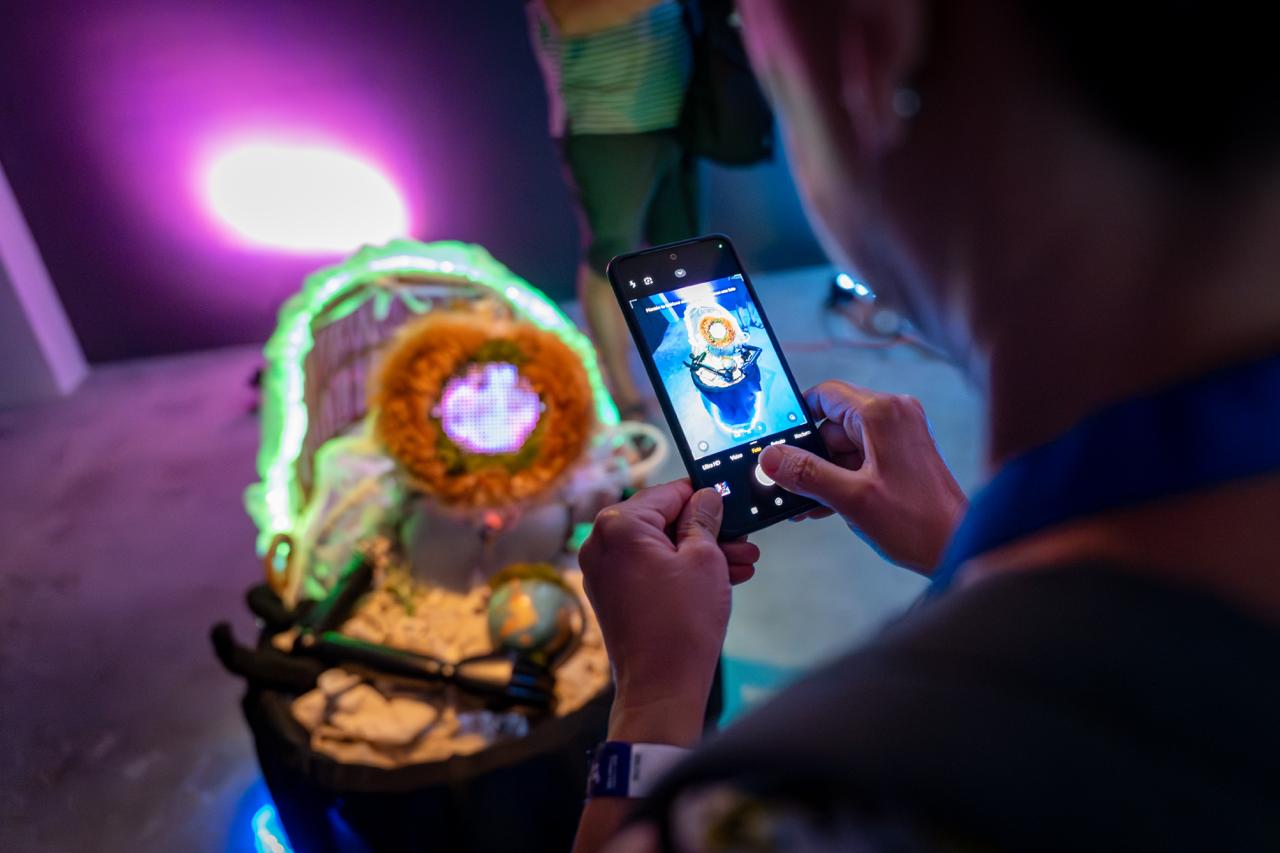 A visitor photographs a lit sculpture of woven textile and orange coral, a small globe resting at its base