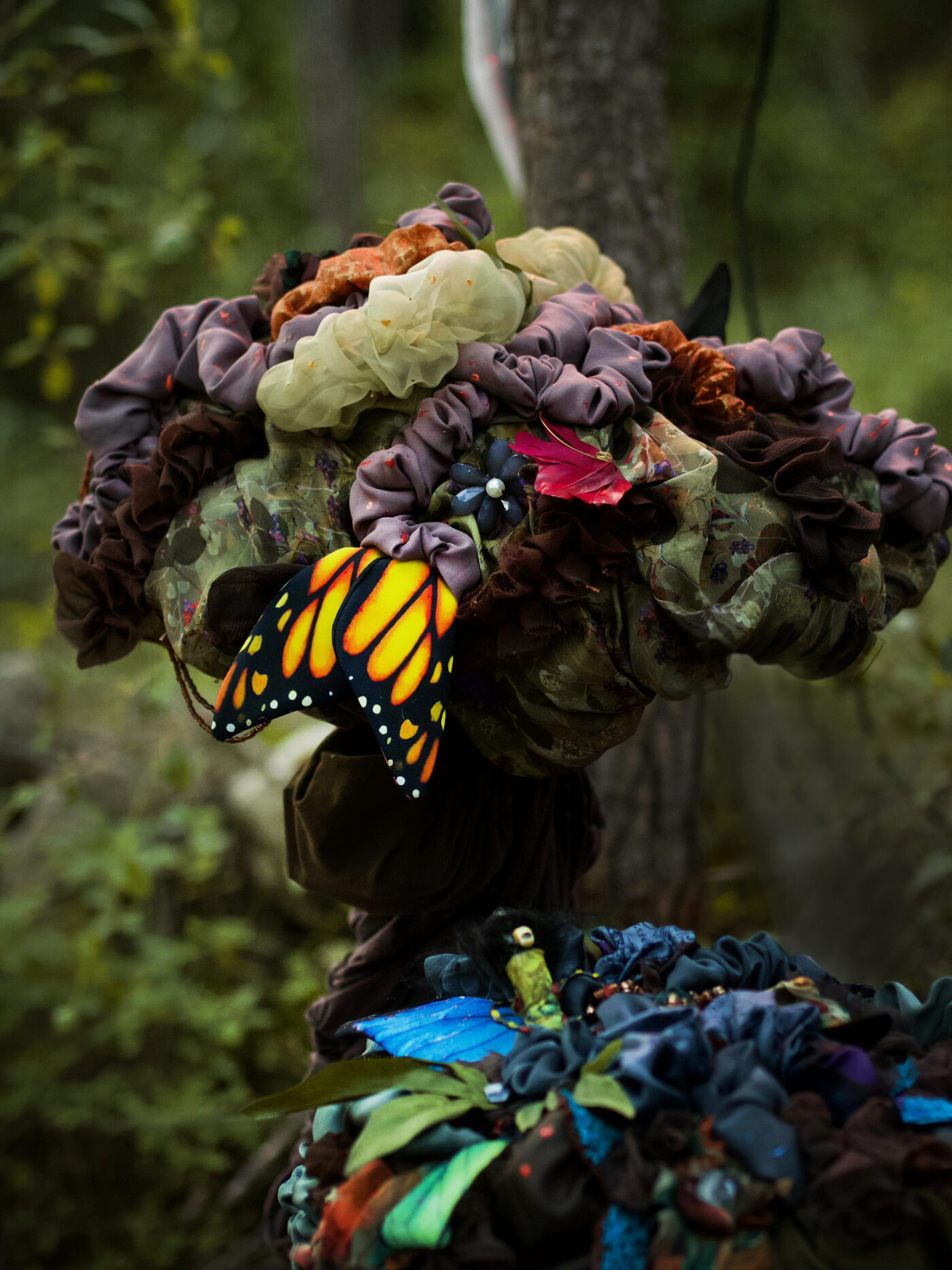 Close view of a cap made from plum, cream and rust fabric rosettes, a large painted monarch wing at the front and a smaller blue and green mushroom behind