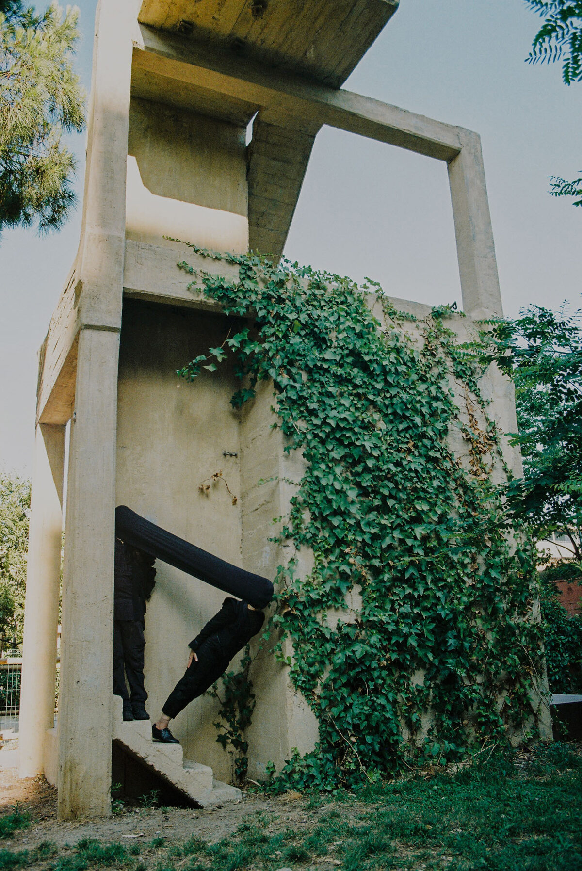 Two figures dressed in black on the steps of an ivy-covered concrete structure, one with a long black sleeve extended overhead