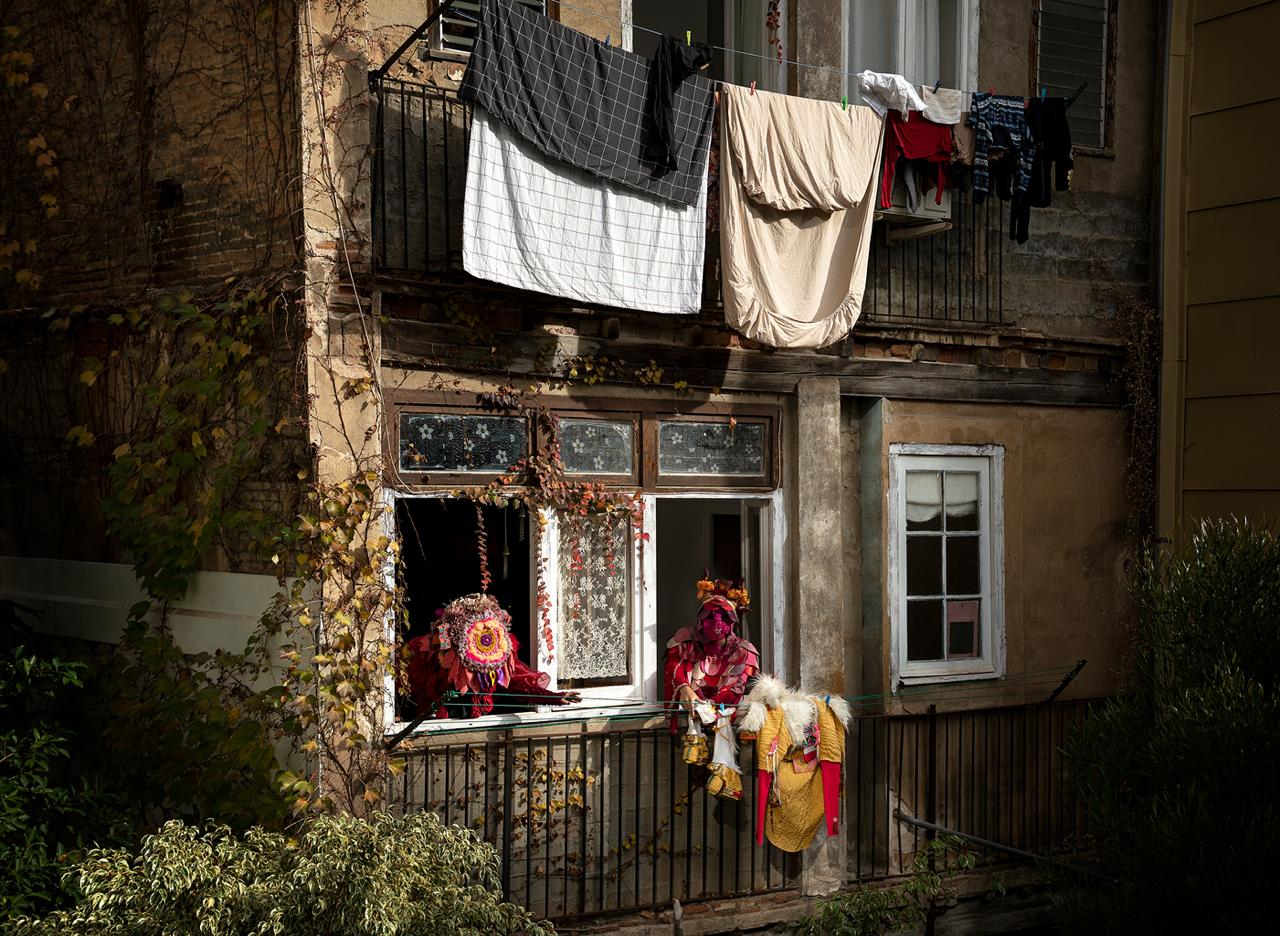 Two costumed figures in red and gold at the lit window of a weathered, ivy-covered building, laundry strung across the balcony above